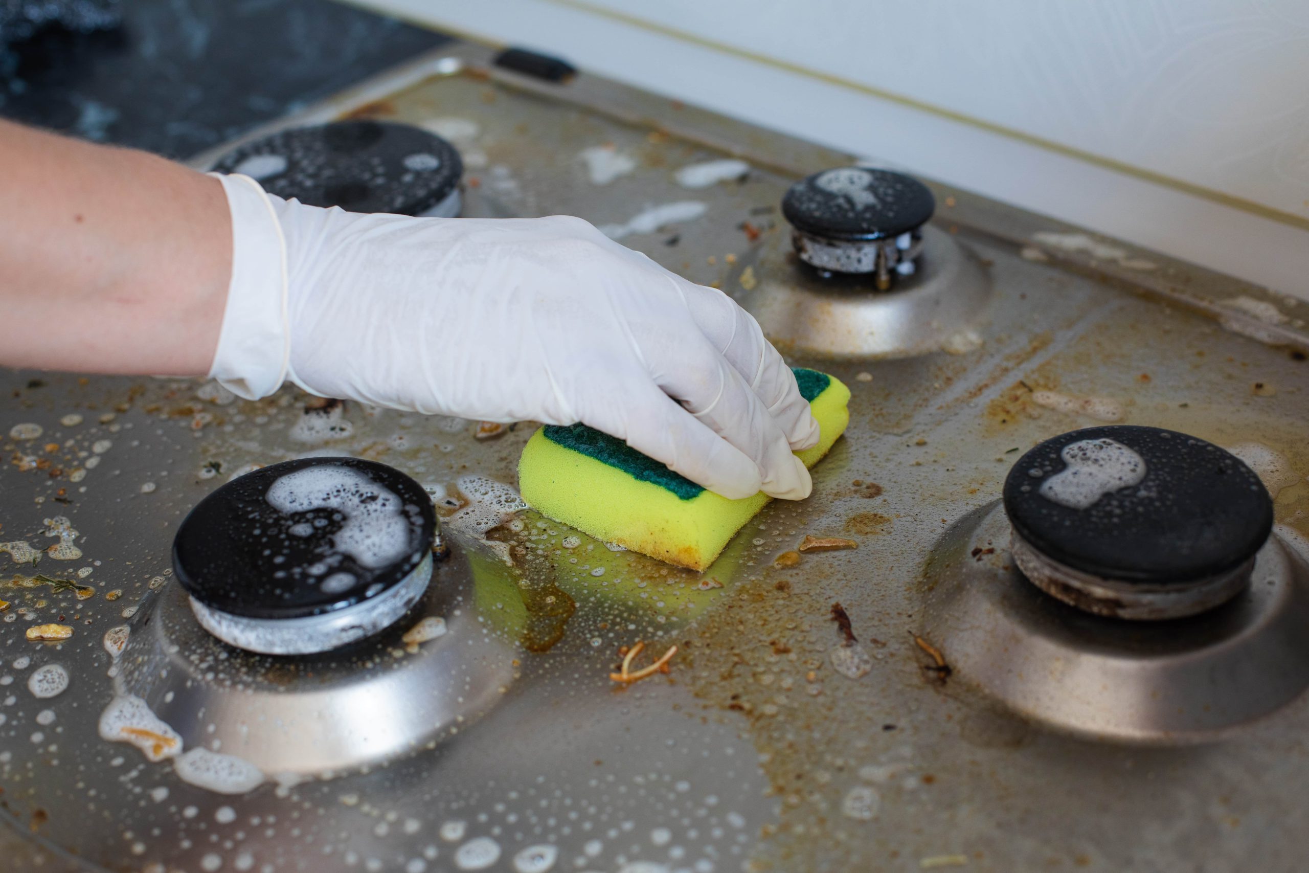Mão feminina doméstica usando luvas limpando um fogão sujo após cozinhar, utilizando uma esponja.