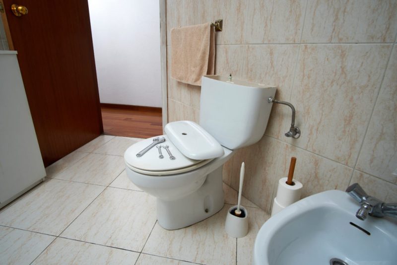 Banheiro com vaso sanitário branco em manutenção, com a tampa da caixa acoplada removida e ferramentas sobre o assento. Ao lado, há pia, porta-papel, escova sanitária, toalha pendurada e parede revestida com azulejos claros